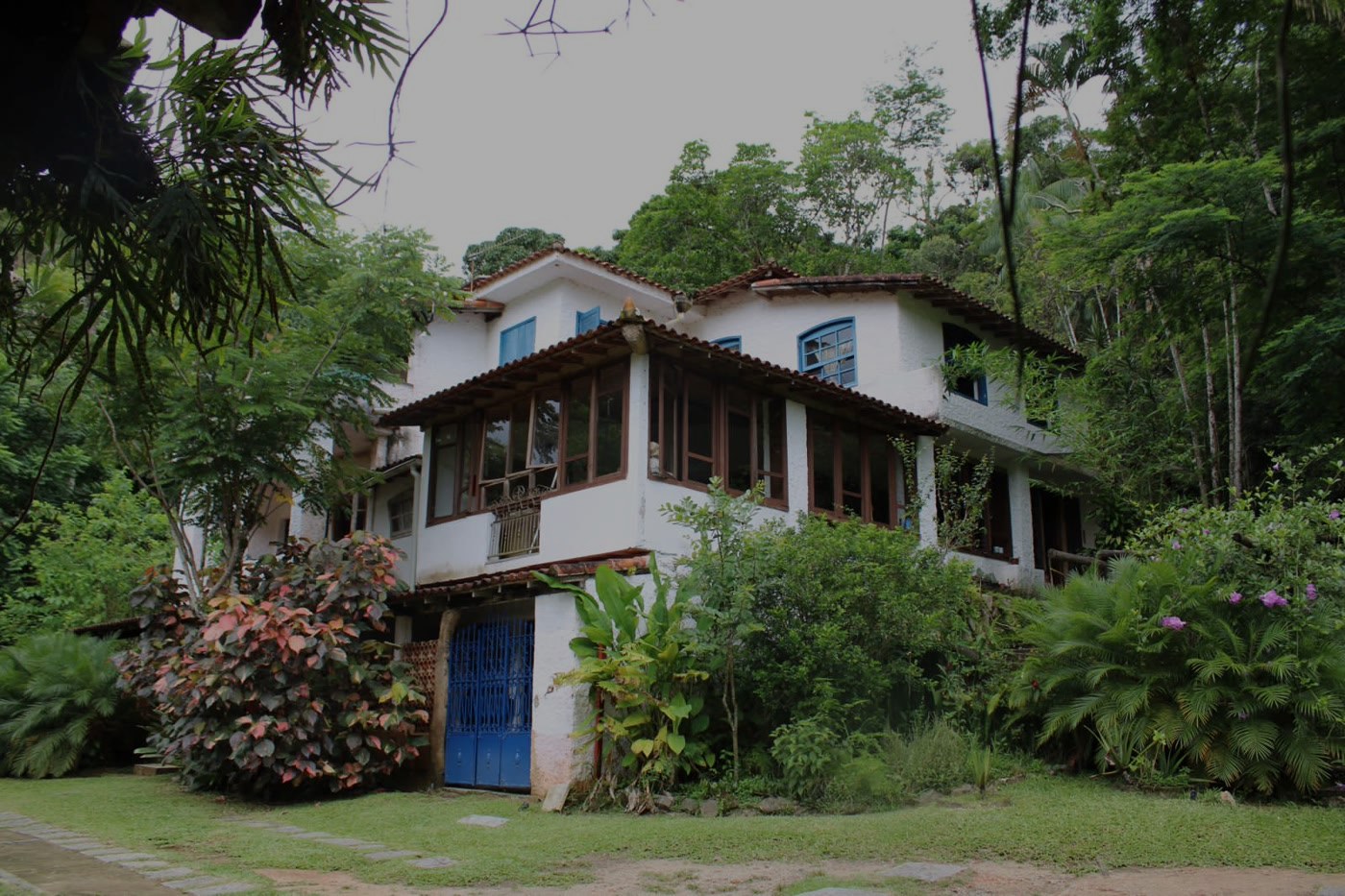 Fachada da casa-sede da Fazenda dos Cordeiros vista do jardim, com varanda de vidro, portão azul e a Mata Atlântica ao redor