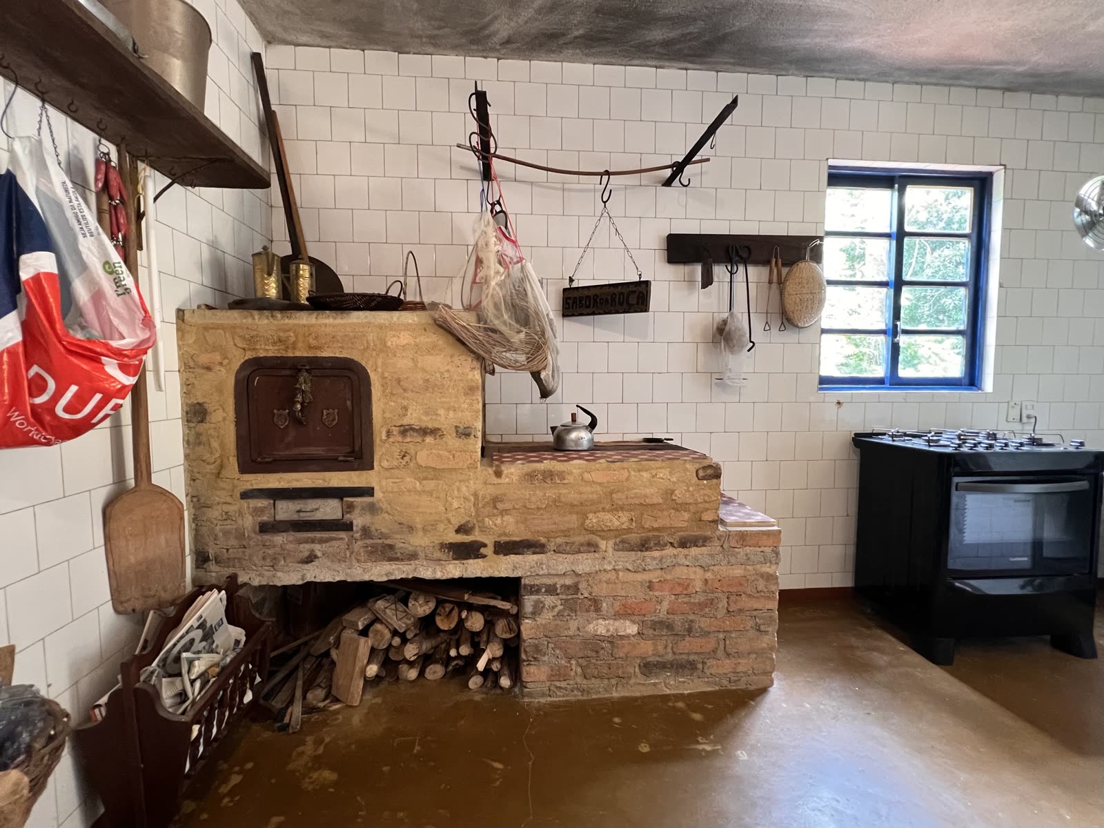 Cozinha da roça com fogão a lenha de tijolo e alvenaria, chaleira sobre a chapa, lenha empilhada embaixo e uma placa de madeira escrita “Sabor da Roça” na parede de azulejo branco