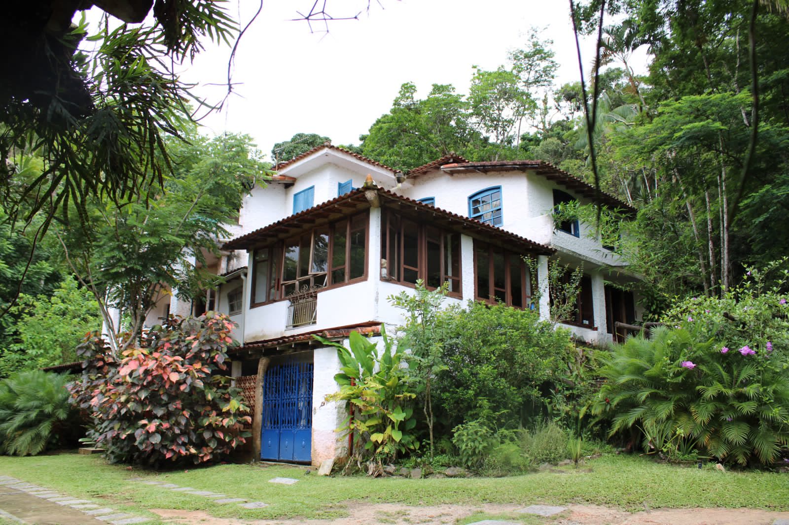 Casa-sede branca de janelas e porta azuis, com arbustos floridos no jardim e a mata alta em volta