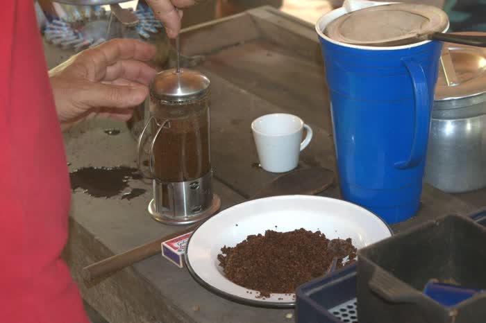 Mãos preparando o café na prensa francesa sobre a bancada de madeira, com o pó moído num prato ao lado