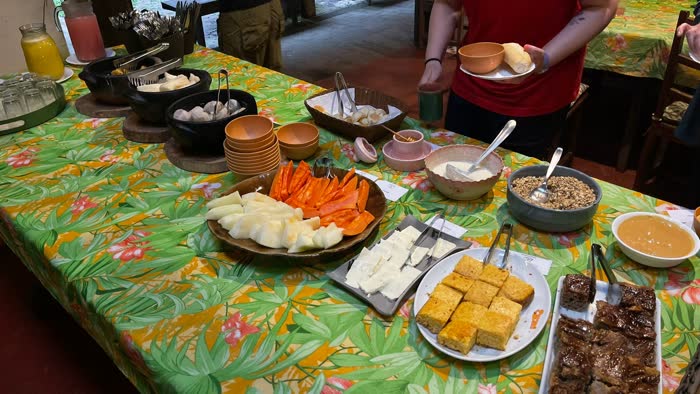 Mesa comprida do café da manhã da fazenda com travessas de melão e mamão, queijo fresco, bolo de fubá, granola e iogurte, e alguém se servindo ao fundo